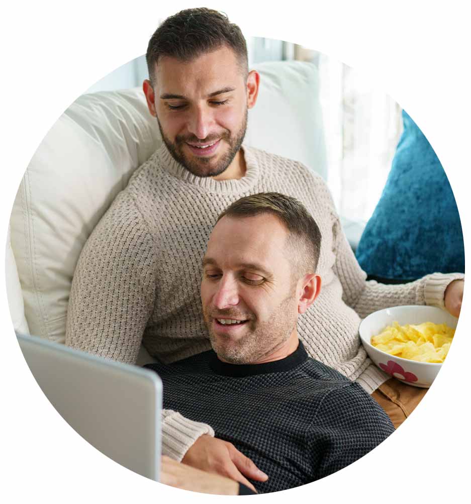 Two men on the couch smiling and watching something on the laptop.