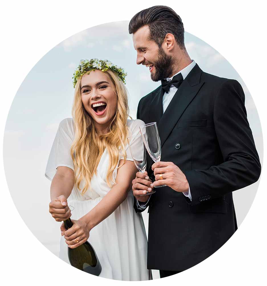 Bride opening bottle of champagne with groom holding glasses.